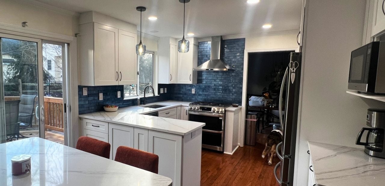 After: Two-tone white and gray Shaker kitchen in Olney with Calacatta quartz, blue glass backsplash, movable island, and stainless chimney hood delivering more storage.